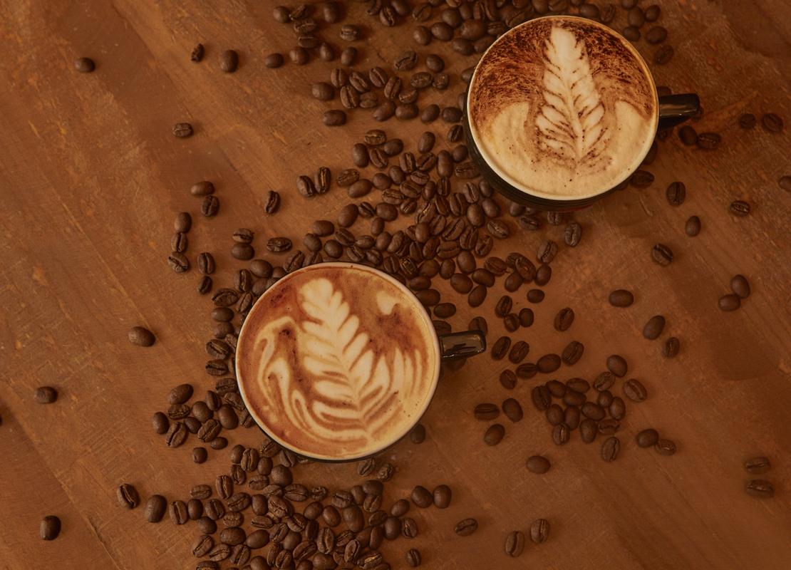 Dos tazas de café con diseño latte art rodeadas de granos tostados sobre una mesa de madera