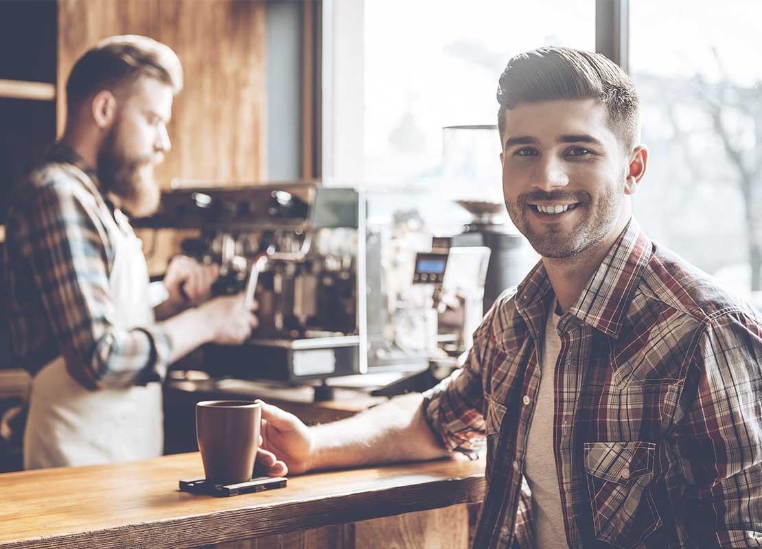 Cliente feliz sostiene una taza y al fondo el barista prepara una bebida utilizando una máquina de café