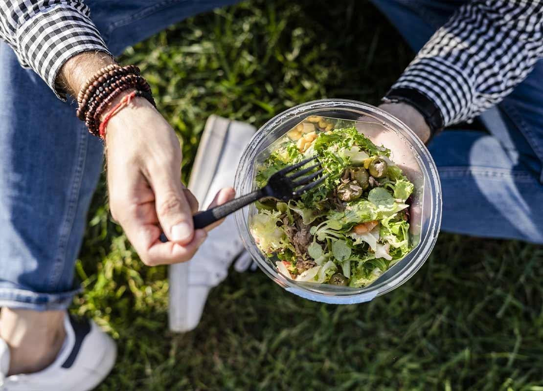 Hombre sentado en el césped sostiene tenedor y ensalada, la cual pidió por un servicio take away