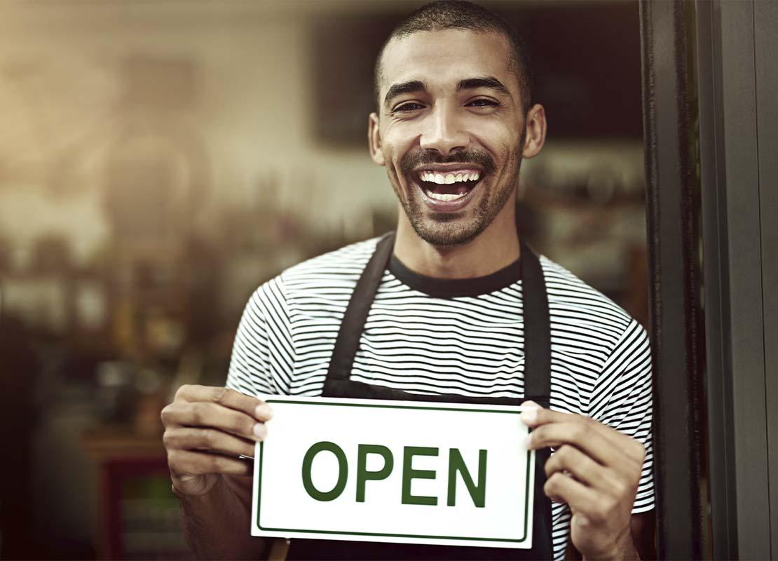 Barista sonriente sostiene en sus manos un cartel de “abierto” en la puerta de una cafetería