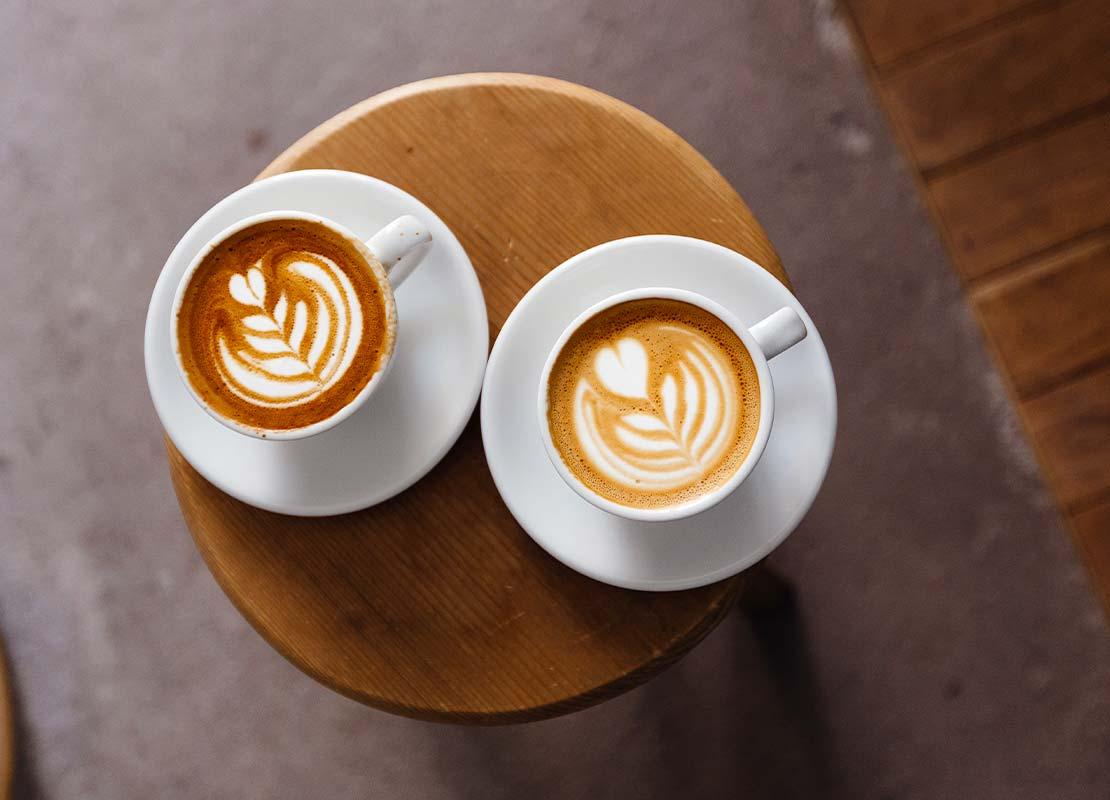 Sobre una mesa, 2 tazas de cappuccino con diseños de latte art con figuras de corazón y hojas