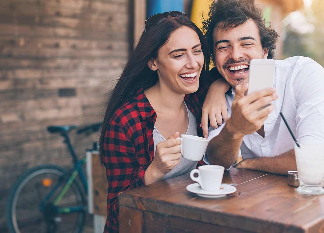 Pareja sentada a la mesa con tazas de café fuera de un restaurante sonríe mientras observa un smartphone