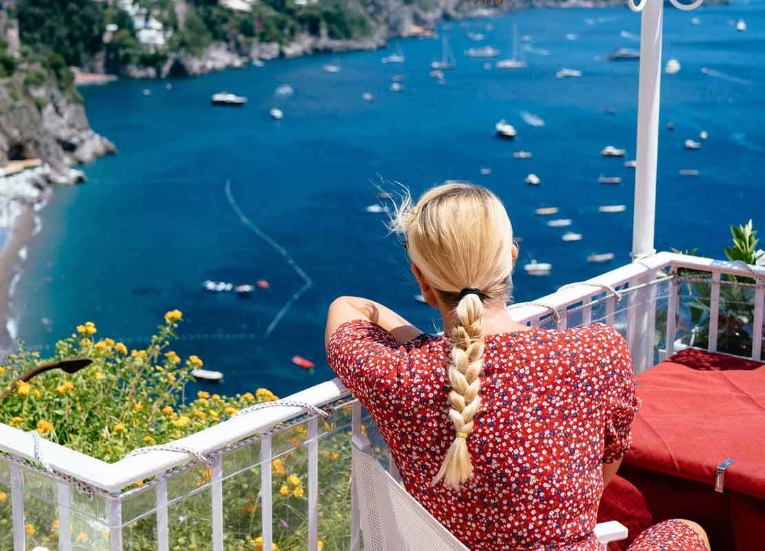 Mujer sentada a la mesa mira desde la terraza de un sky bar el mar, la playa y los botes a lo lejos