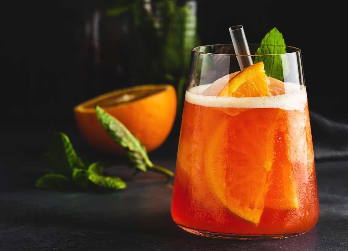 Vaso de cristal con cóctel sin alcohol de naranja y hierbabuena, al fondo la fruta y las hierbas aromáticas