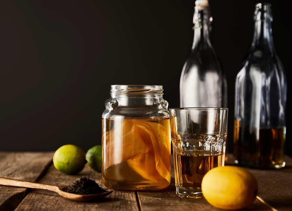 Frasco y vaso de cristal con kombucha, al lado limones, una cuchara de madera y más botellas altas