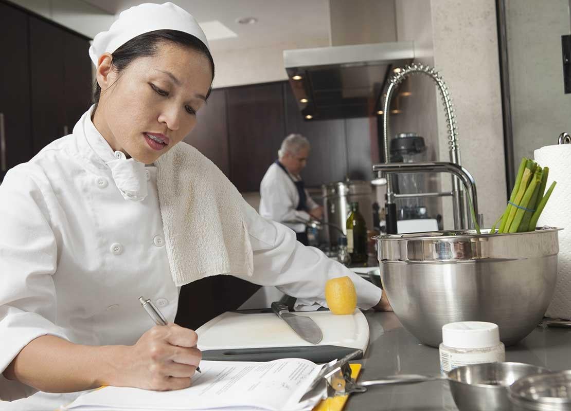Chef escribe en una planilla sobre un mesón de cocina comercial mientras manipula utensilios