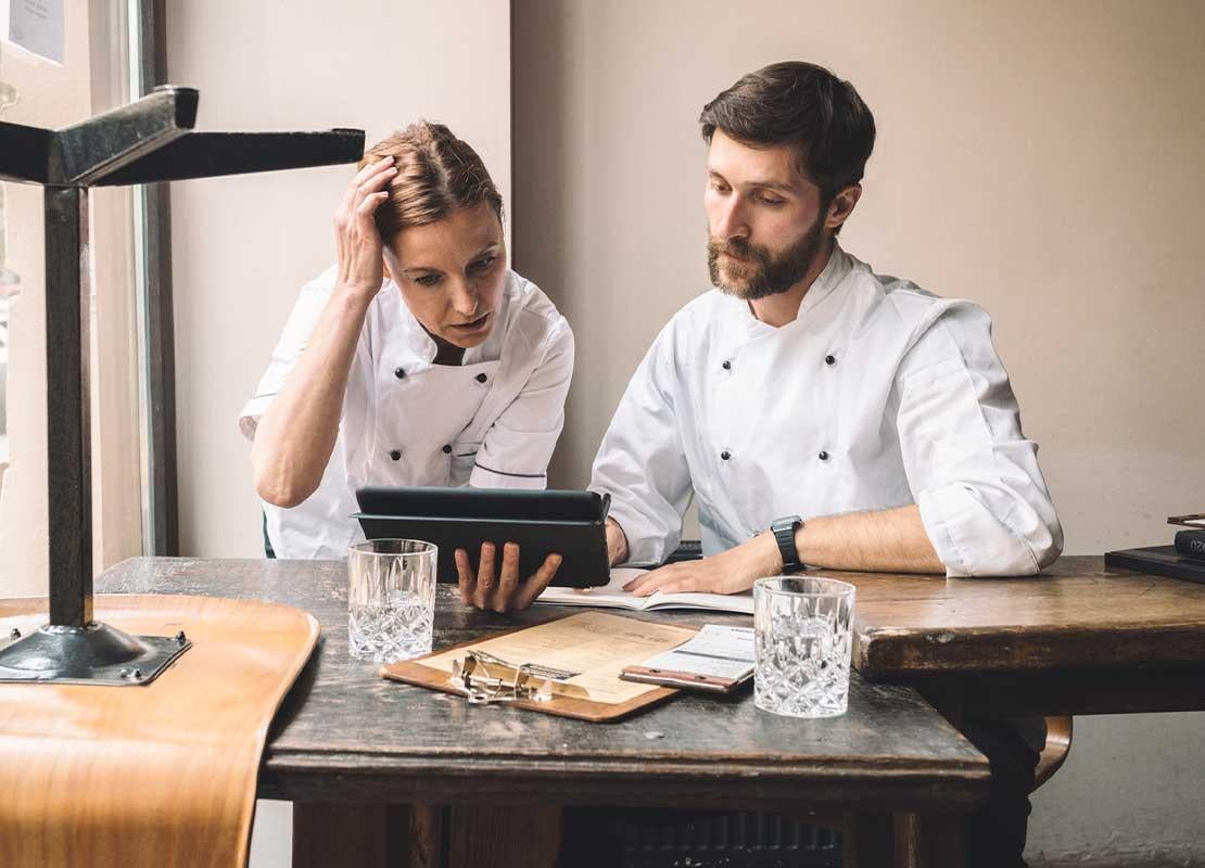Dos chefs revisan una tablet, sentados a la mesa de una oficina sobre la que hay vasos