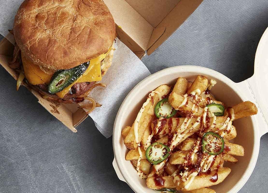 Una hamburguesa servida en una caja de comida y papas con salsa en un recipiente circular reciclable