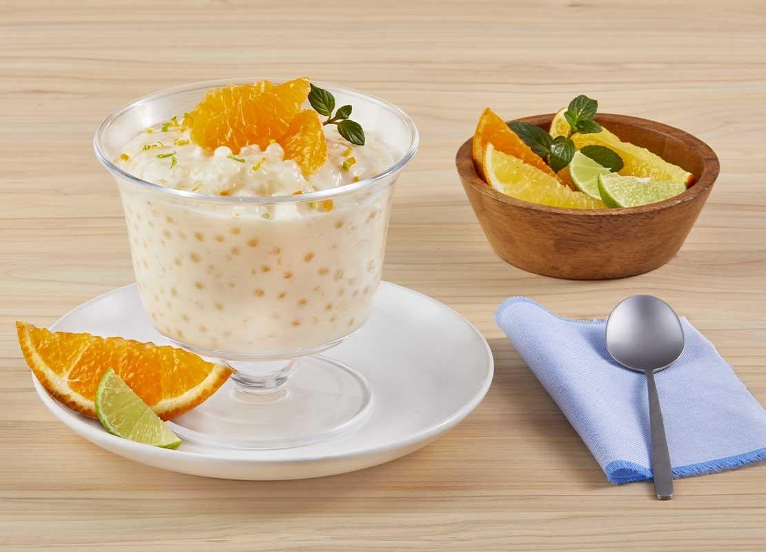 Copa de tapioca con leche condensada, coronada con naranja en gajos, al lado una taza con frutos cítricos