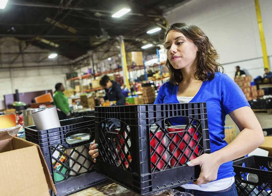 En una fábrica, mujer sostiene una caja con productos, al fondo, otros trabajadores, estanterías y cajas