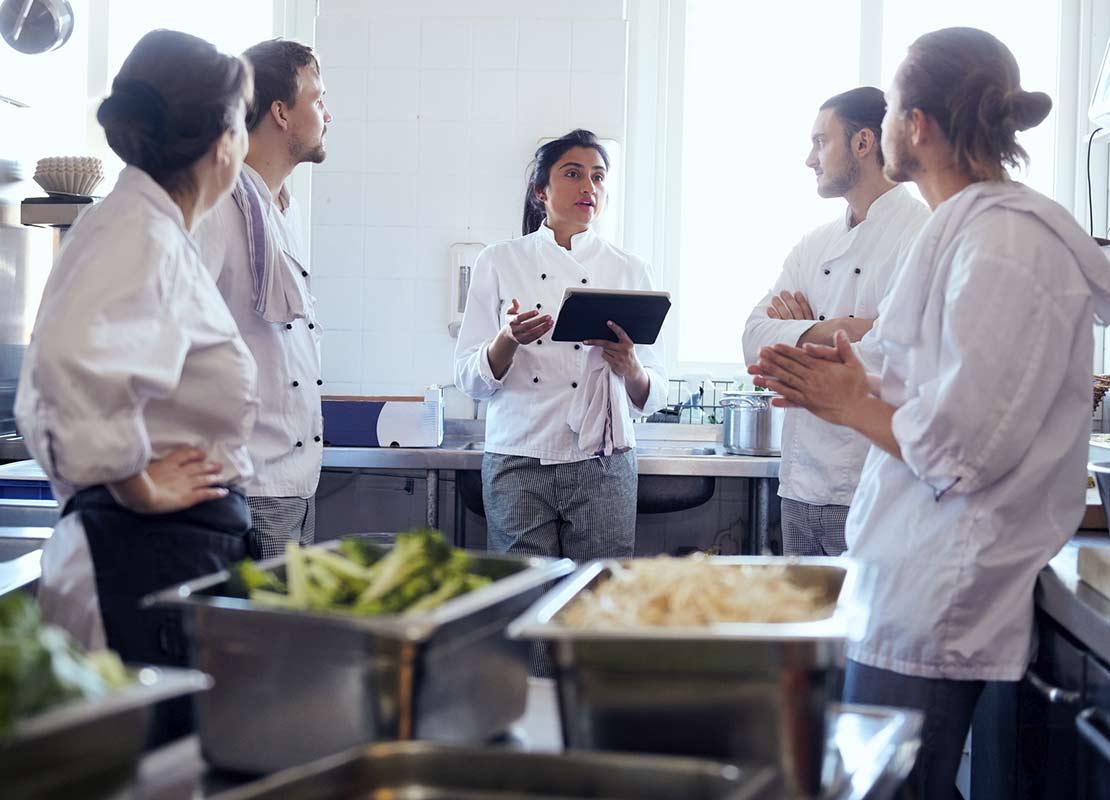 En una cocina comercial, chef sostiene una tablet y explica a 4 cocineros para capacitarlos