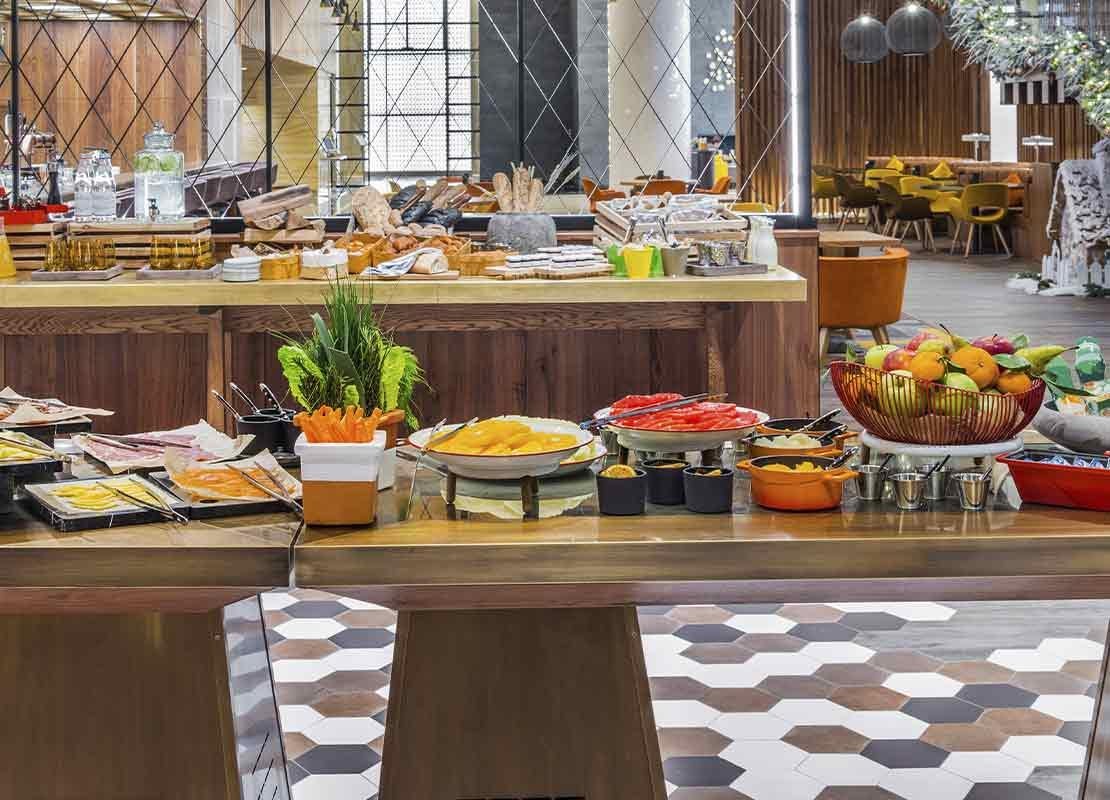 Salón con varias mesas de buffet con diversos alimentos como platos fuertes, ensaladas, bebidas, etc.