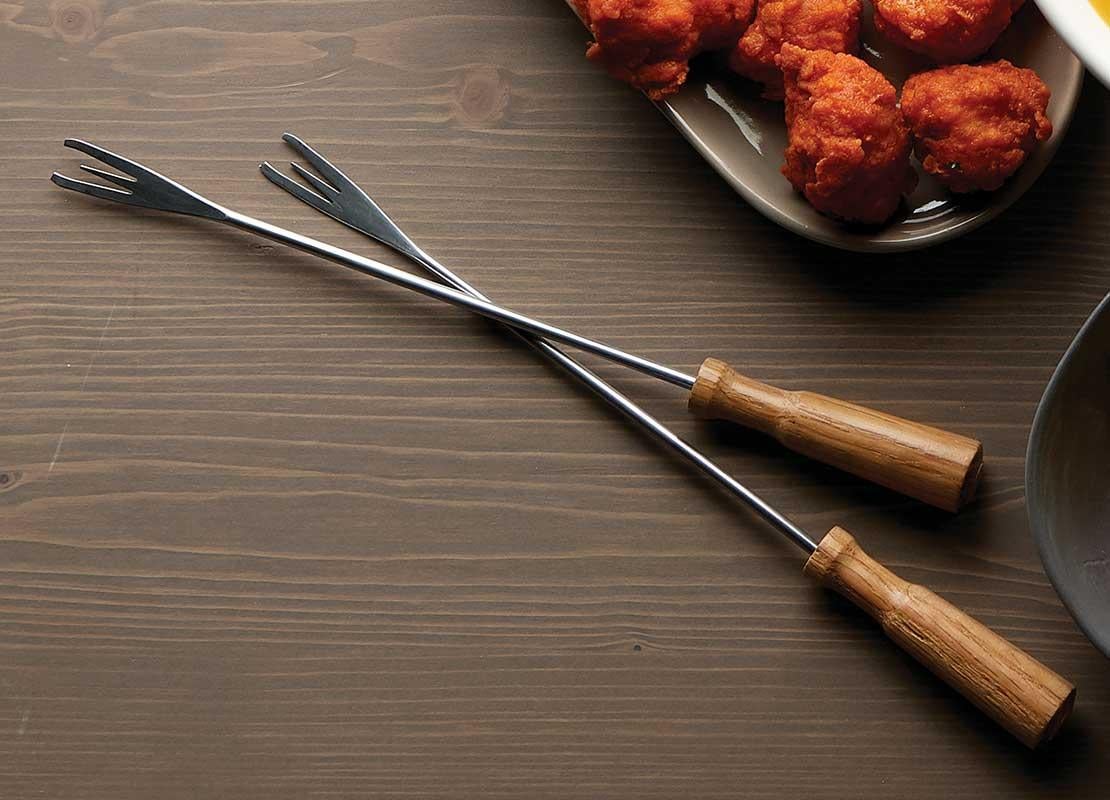 Sobre una mesa dos tenedores para fondue, salsa de queso, Nuggets de pollo y papas artesanales