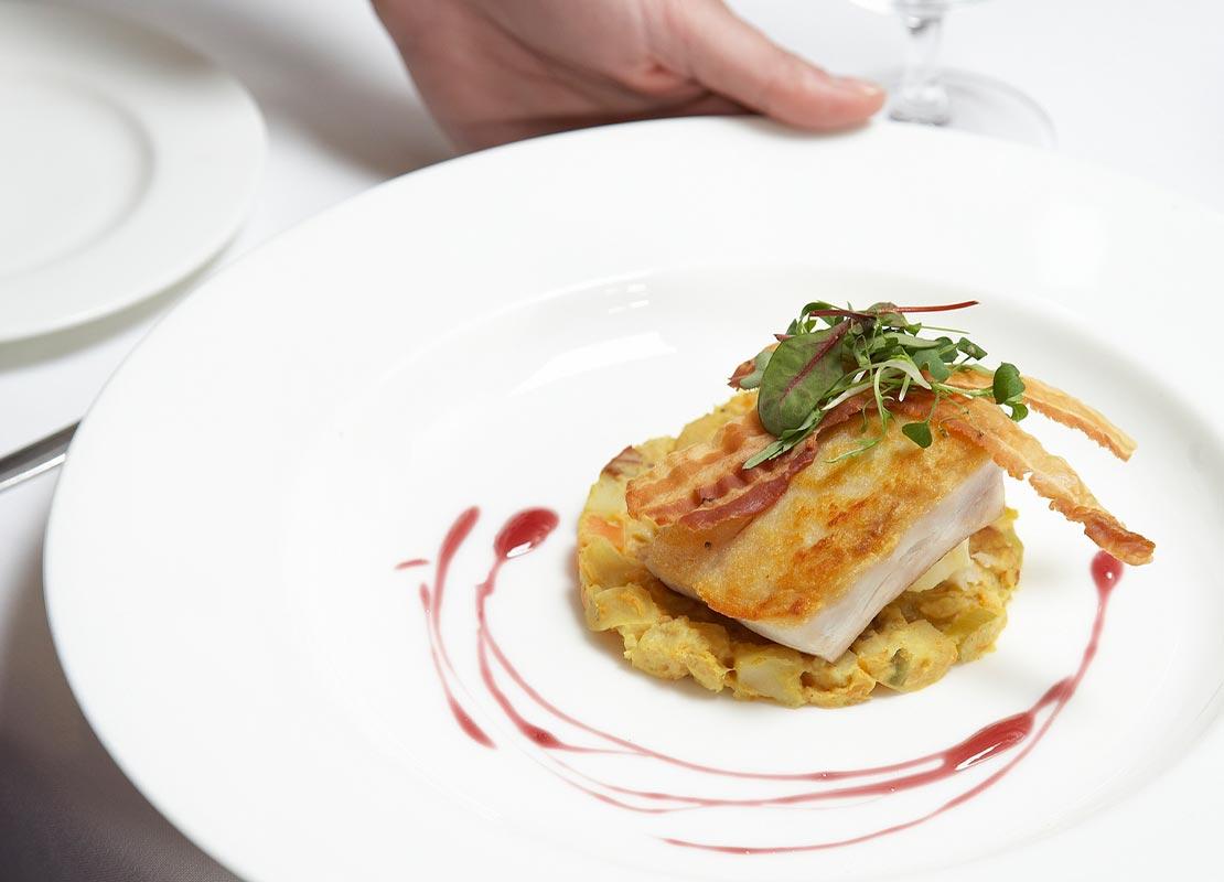 Mano de mesero pone un plato de comida sobre la mesa de un comensal, al lado copas y platos