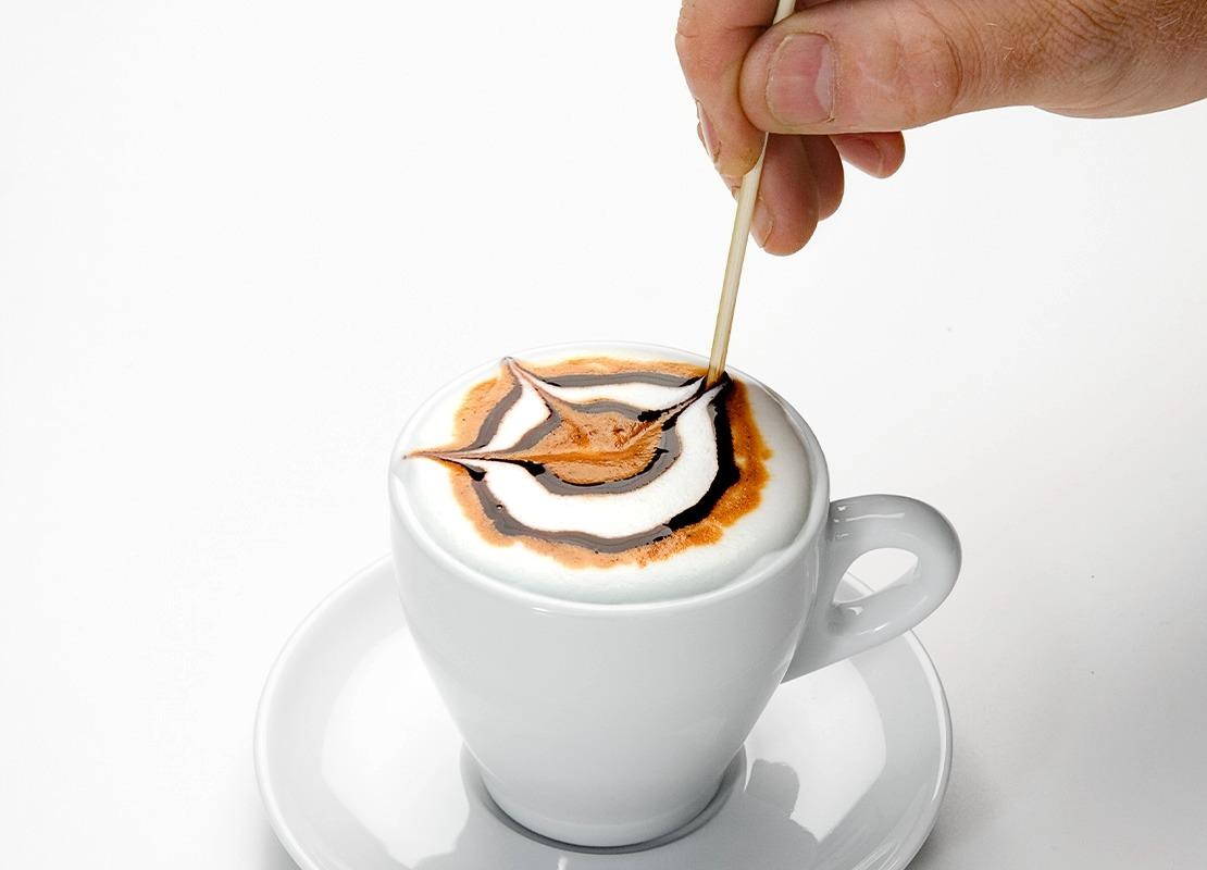 Mano decorando un café con arte latte usando un palillo sobre espuma y jarabe