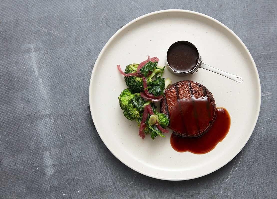 Hamburguesa sin pan bañada en salsa, servida con brócoli, cebolla encurtida y un recipiente con más salsa
