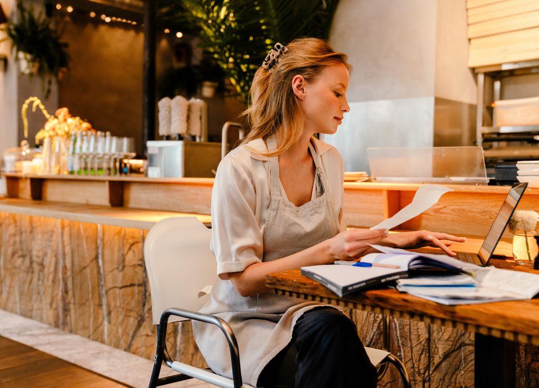 Propietaria de restaurante revisa documentos físicos junto a su laptop en la barra del local.