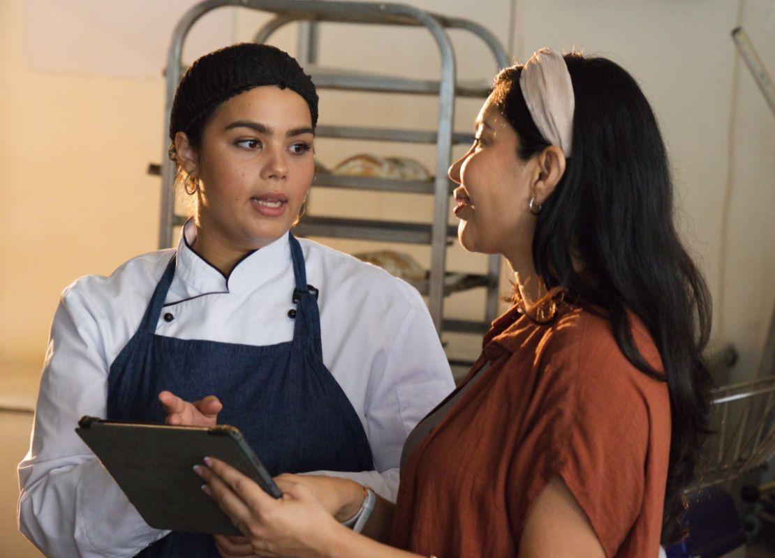 Chef en delantal azul habla con una supervisora mientras revisan una tableta en la cocina.