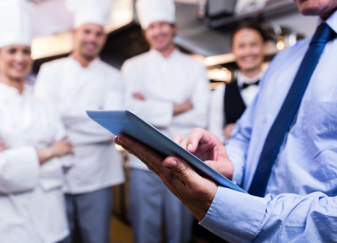 Gerente en camisa azul revisa una tableta frente a su equipo de cocina sonriente al fondo.