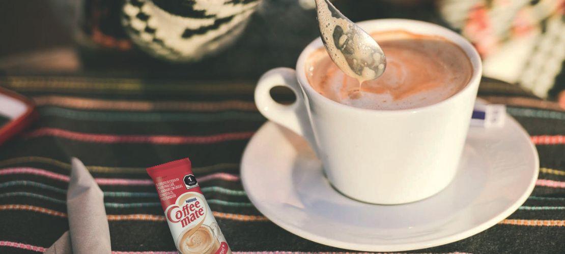 Persona revolviendo café caliente en una taza con cuchara junto a un sobre de sustituto de crema.
