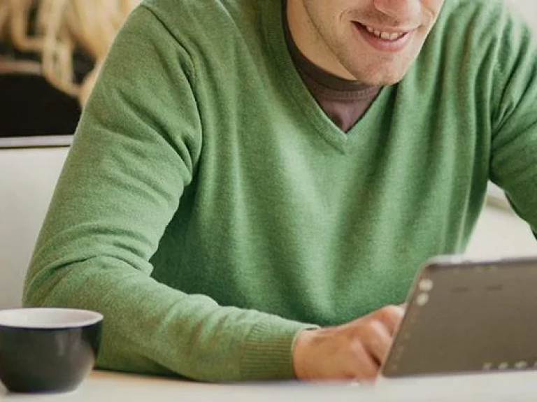Hombre de sueter verde consulta novedades en su tablet mientras disfruta un café