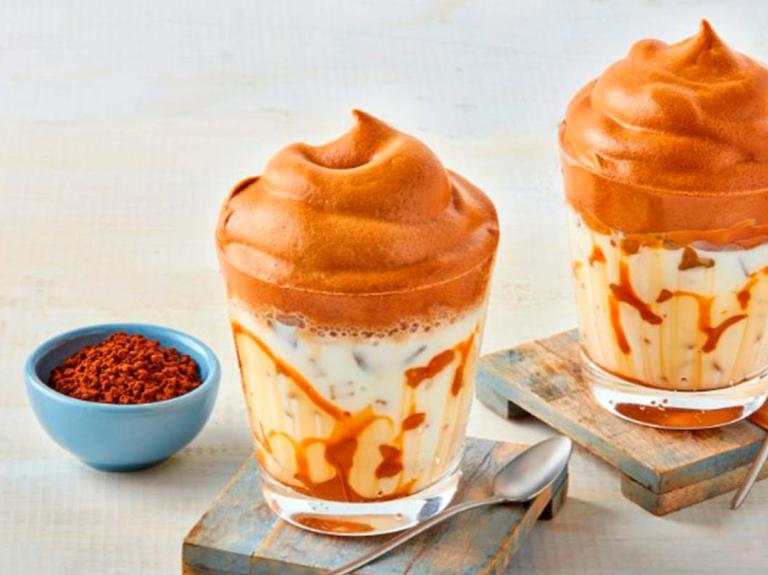 Dos vasos de café frío con hielo, leche y coronados con crema de café sobre portavasos de madera y junto dos cucharas