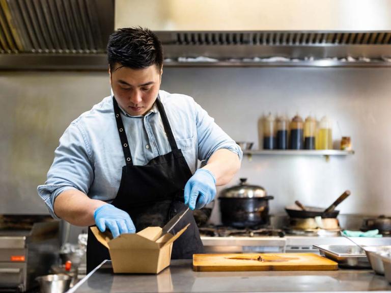 En una cocina comercial, chef empaca una preparación en una caja para ser enviada como entrega a domicilio