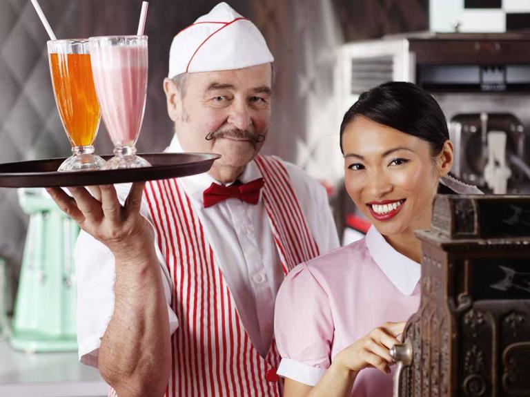 En restaurante temático de los 50, un mesero sostiene una bandeja con jugos y otra opera la caja registradora