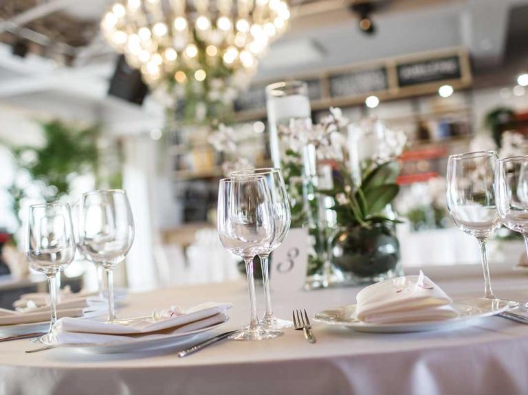 Juego de mesa con copas, platos blancos y lámparas de araña de luz en el fondo que aportan a la decoración de restaurantes