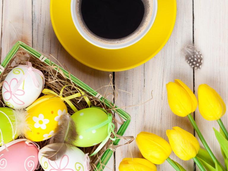 Sobre una mesa de madera 5 rosas amarillas, una taza de café y una canasta con dulces de pascua como huevos decorados