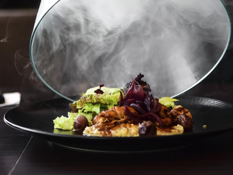 Sobre una mesa de madera se destapa un plato de comida gourmet envuelta en humo