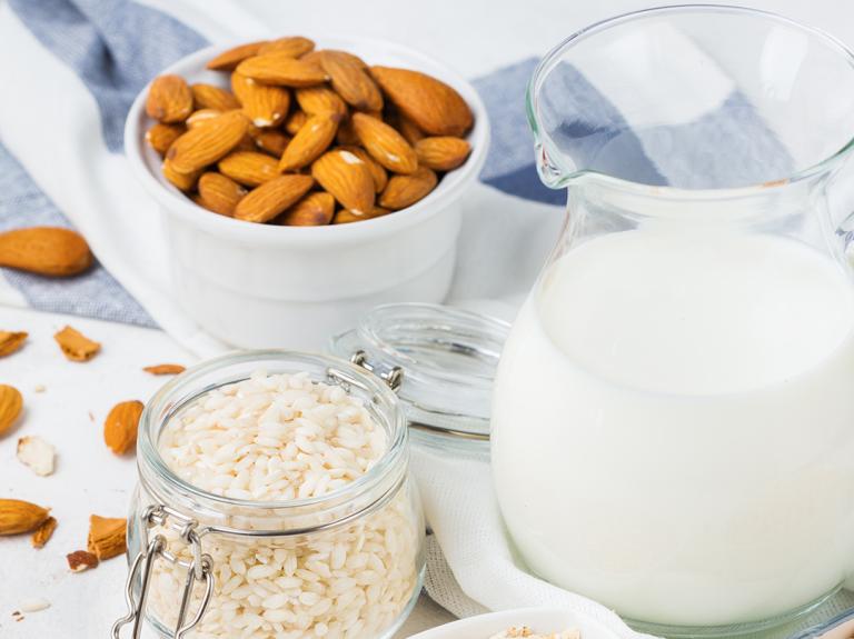 Sobre una mesa una jarra con una bebida vegetal y a su alrededor recipientes con avena, arroz, almendras y garbanzos