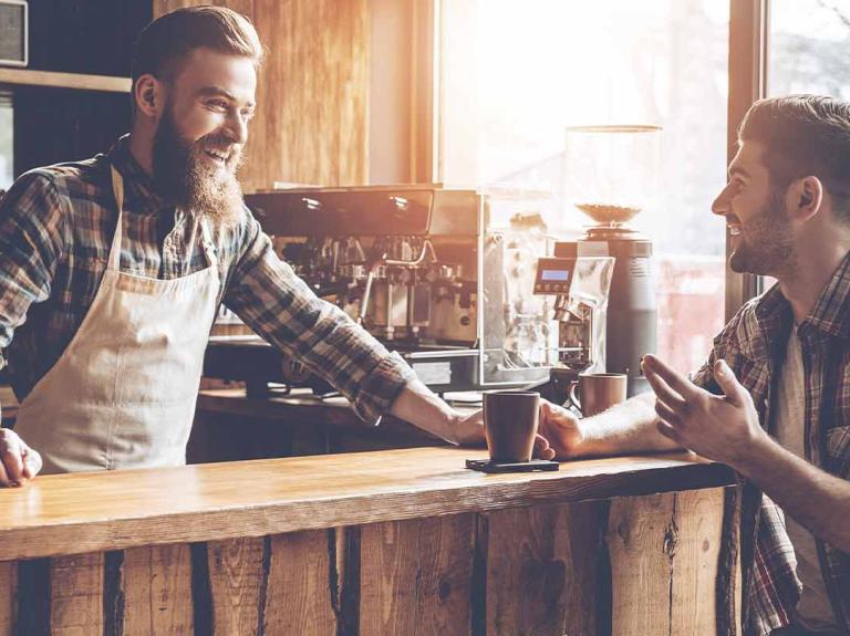 Barista atiende en su cafetería a un cliente sentado a la barra que sostiene una taza de café 