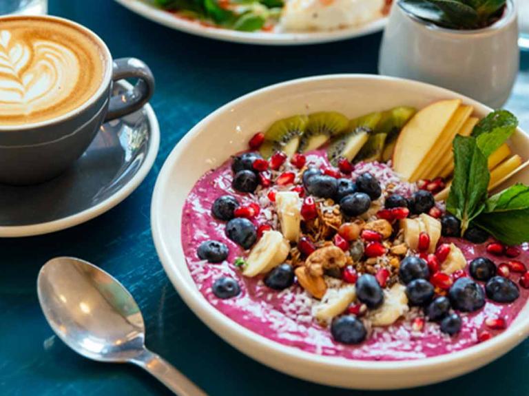 Servido a la mesa un bowl con yogurt y fruta, cappuccino y vasos de agua, que componen un desayuno