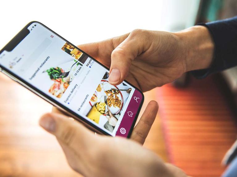 Manos sostienen un celular que muestra una plataforma de entrega a domicilio con diferentes platillos