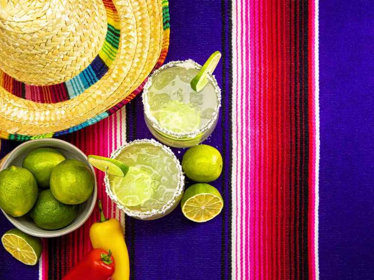 Dos vasos de michelada con rodajas de limón en el borde y al lado la fruta, un sombrero y maracas