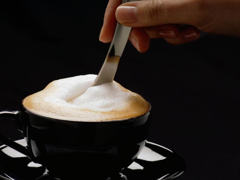 Mano de una mujer mezcla con una cuchara un cappuccino NESCAFÉ® servido en una taza negra
