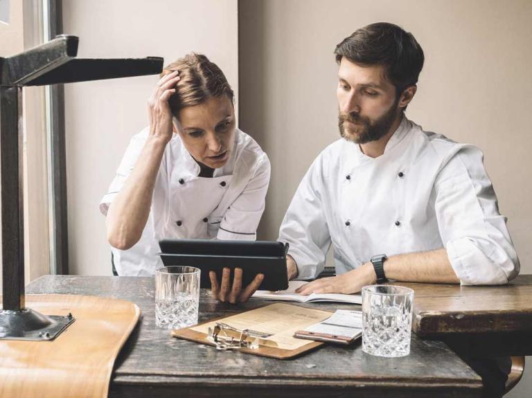 Dos chefs observan una tablet sentados a la mesa donde hay vasos, libretas y tabla legajadora