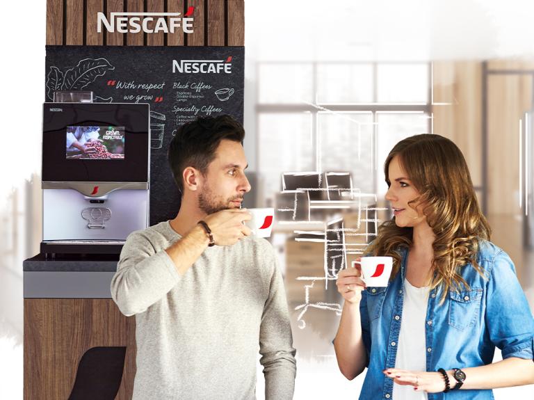 Dos compañeros de trabajo disfrutan de una taza de café en la oficina, al fondo una máquina NESCAFÉ®