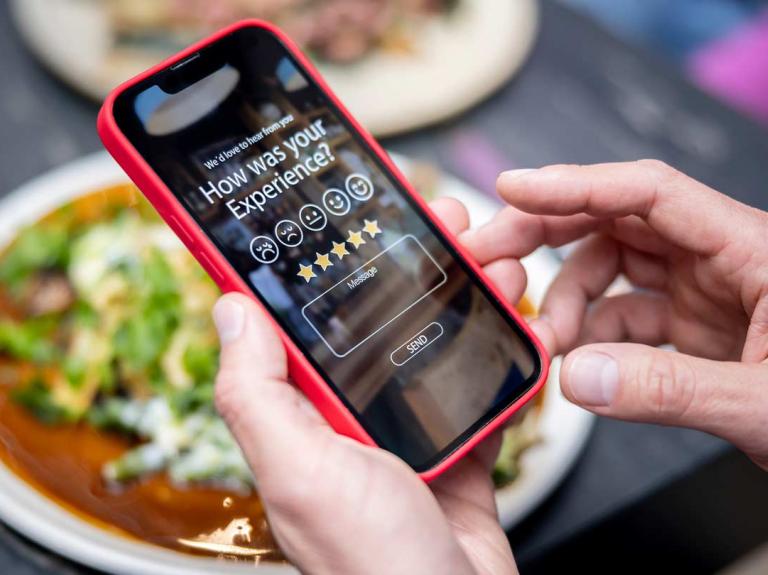 Manos de hombre sostienen un smartphone donde califica su experiencia en un restaurante frente a un platillo