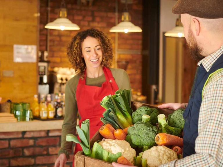 Dueña de un negocio recibe a su proveedor de alimentos local quien lleva una caja con vegetales frescos