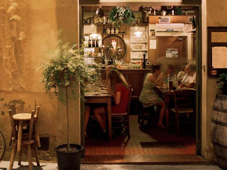 Fachada de una hostería con comensales al fondo sentados a la mesa y afuera sillas, plantas y bicicleta