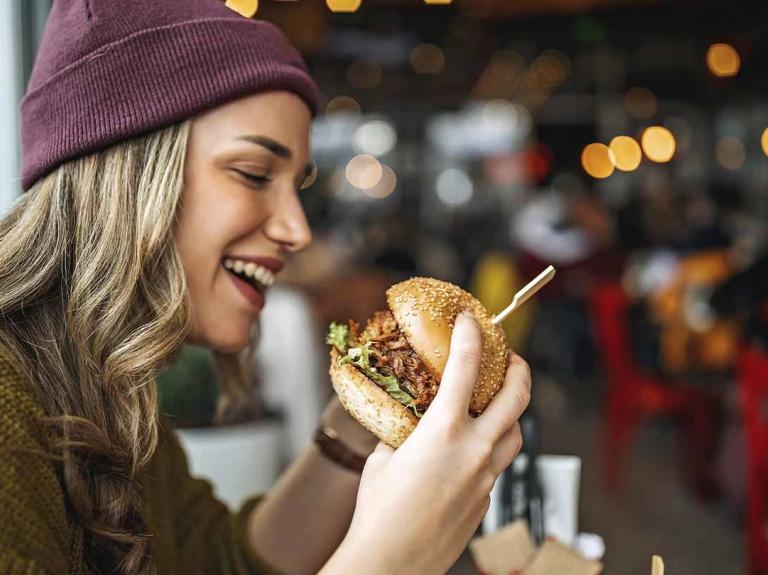 Mujer observa una hamburguesa que sostiene en sus manos, al lado bebida y otros apretivios