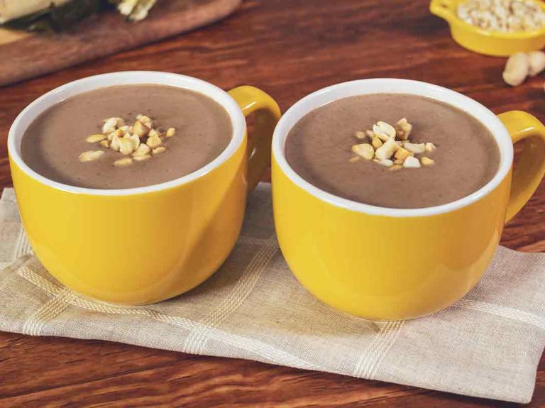 Dos tazas amarillas de chocolate caliente coronadas con cacahuates, al fondo hallacas sobre una tabla