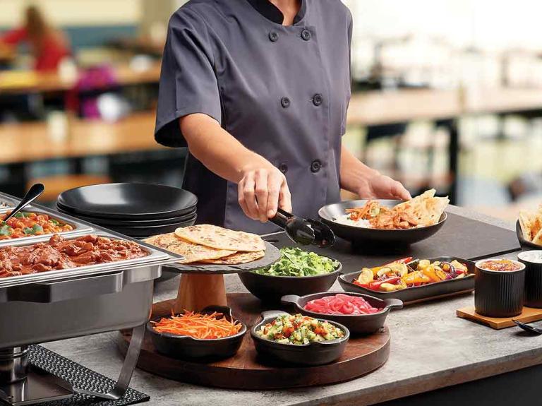 Chef frente de una mesa de buffet sostiene unas pizas para tomar la ensalada de una taza