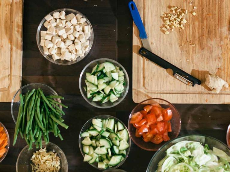 Mesa con tablas de cortar, tazas con alimentos cortados de distintas formas, alrededor, manos de mujeres