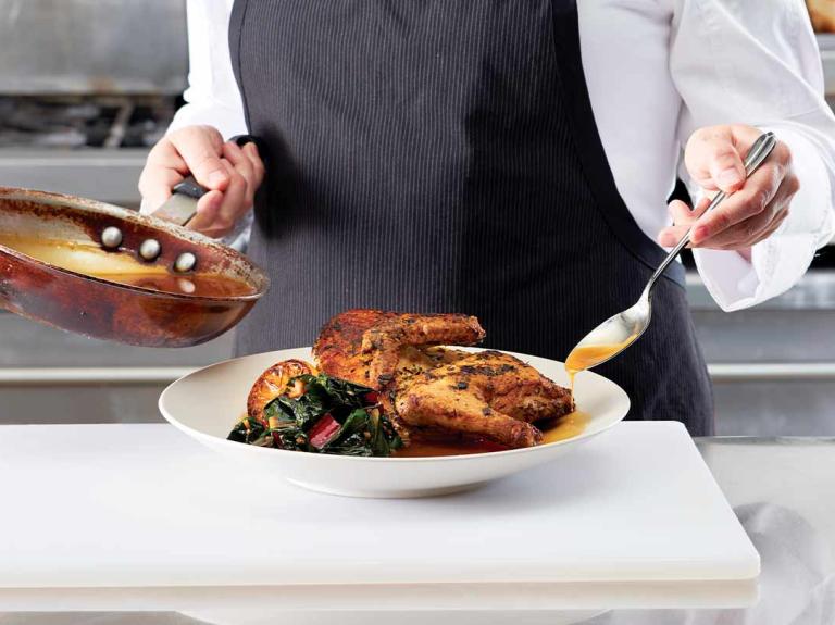 Chef adereza pollo servido en un plato sobre el mesón de una cocina, en otra mano tiene una sartén