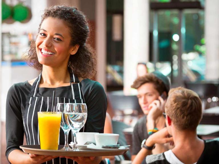 Mesera sonríe mientras sostiene una bandeja con copas, tazas y jugo, al fondo comensales sentados