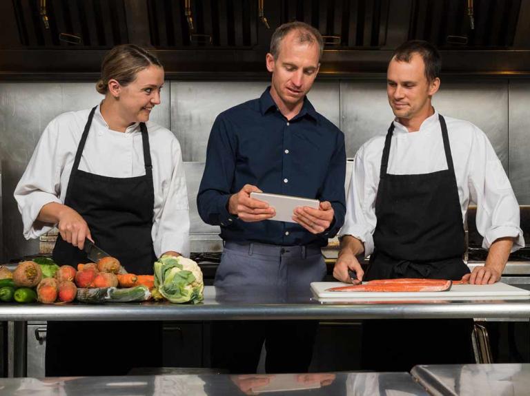 Dos chefs y un gerente de cocina revisan una tablet, rodeados de ingredientes frescos en una cocina profesional