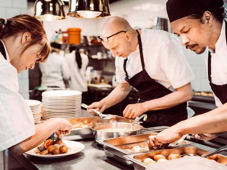 3 chefs en una cocina comercial seleccionan alimentos de la mesa para emplatar las preparaciones de sus clientes